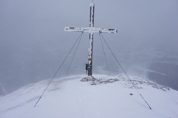 Beim Gipfelkreuz peitscht böiger Wind scharfe Schneekörner ins Gesicht